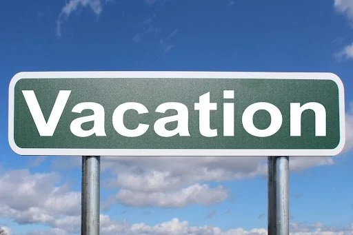 Green road sign with the word 'Vacation' in white letters, set against a cloudy blue sky.
