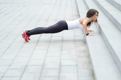 Woman in white tank top and black leggings doing a push-up on a paved outdoor surface.
