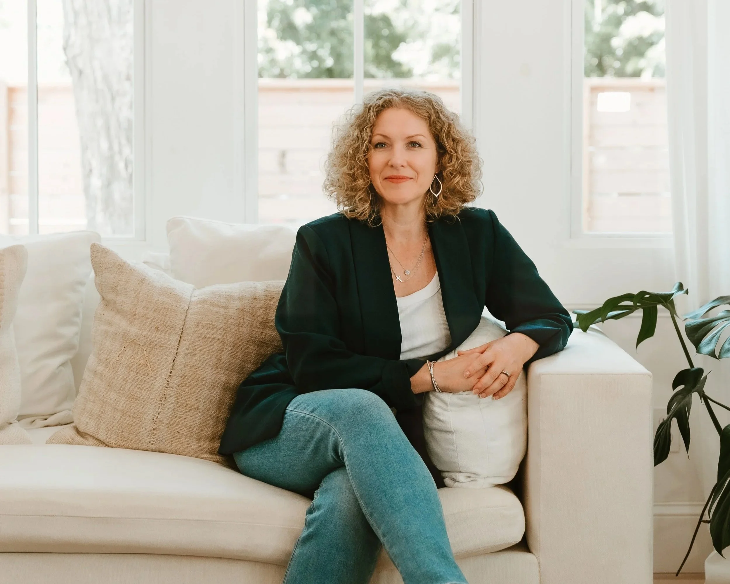 A woman with curly blonde hair sitting on a cream-colored sofa in a bright room with large windows and a wooden fence outside. She is wearing a black blazer, white top, and blue jeans, with her hands resting on a pillow.