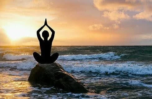 Silhouette of a person practicing yoga on a rock in the ocean during sunset.