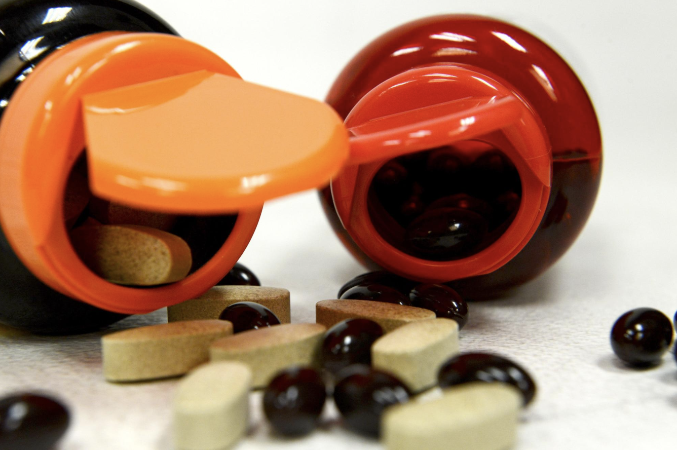An overturned orange and red pill bottle with black jelly beans and various pills spilled onto a white surface.