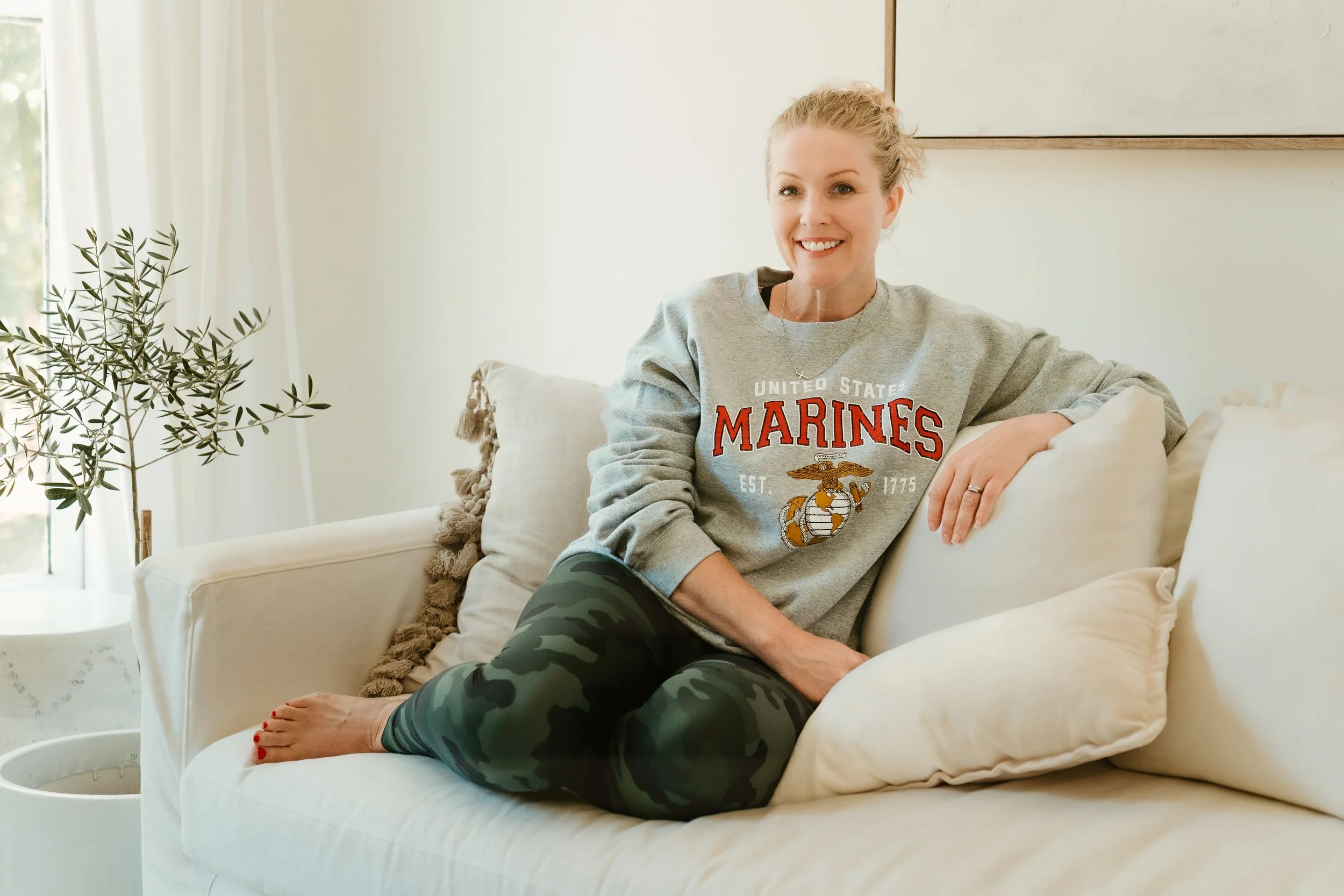 A woman sitting on a light-colored sofa in a bright room, smiling at the camera. She is wearing a gray United States Marines sweatshirt and camouflage leggings, with her legs crossed on the sofa. There is a potted plant and curtains in the background.