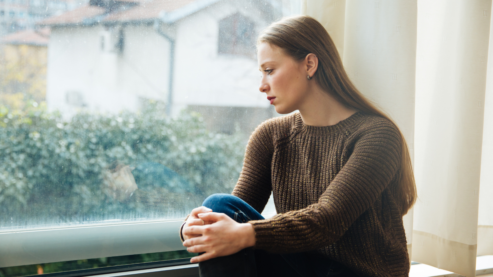 A woman sitting on a window sill looking outside on a rainy day, with a pensive expression.
