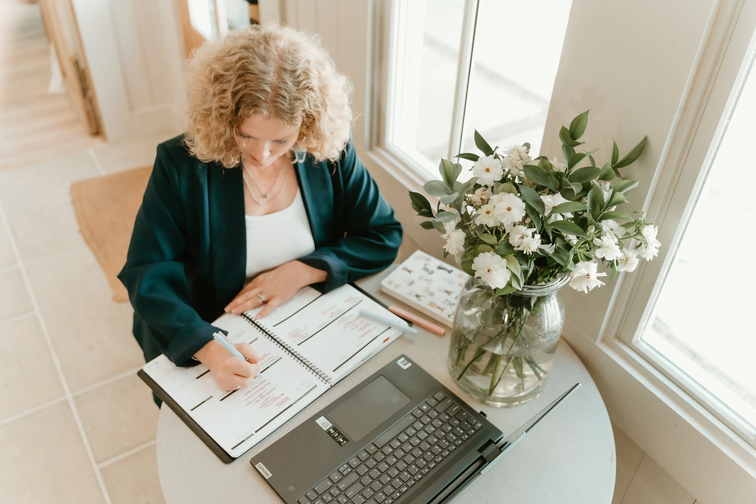 A woman with curly blonde hair sitting at a round table, writing in a planner, with a laptop and a large vase of white flowers on the table, near a window.