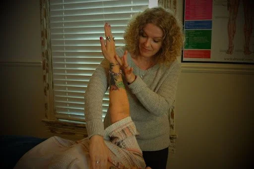 A woman receiving physical therapy on her arm in a clinic room with blinds and anatomical posters on the wall.