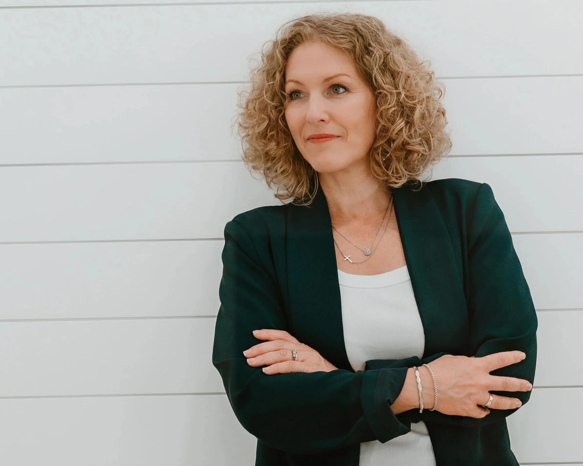 A woman with curly blonde hair standing against a white wall, wearing a black blazer over a white top, with necklaces and bracelets, arms crossed.