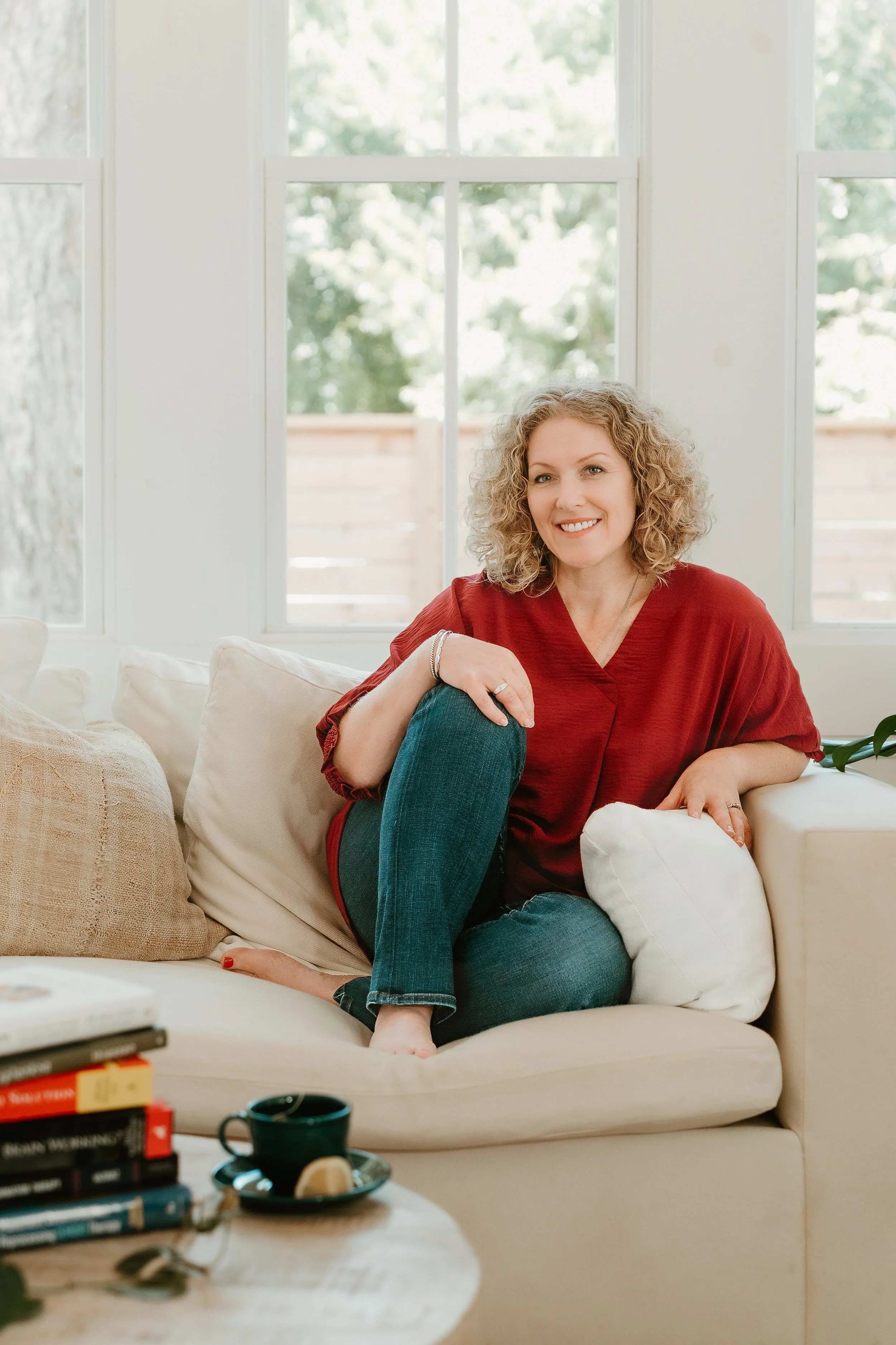 A woman with curly blonde hair sitting on a cream-colored couch, wearing a red top and jeans, smiling. A window with greenery outside is behind her, and there are books, a cup, and pillows on the couch and table.