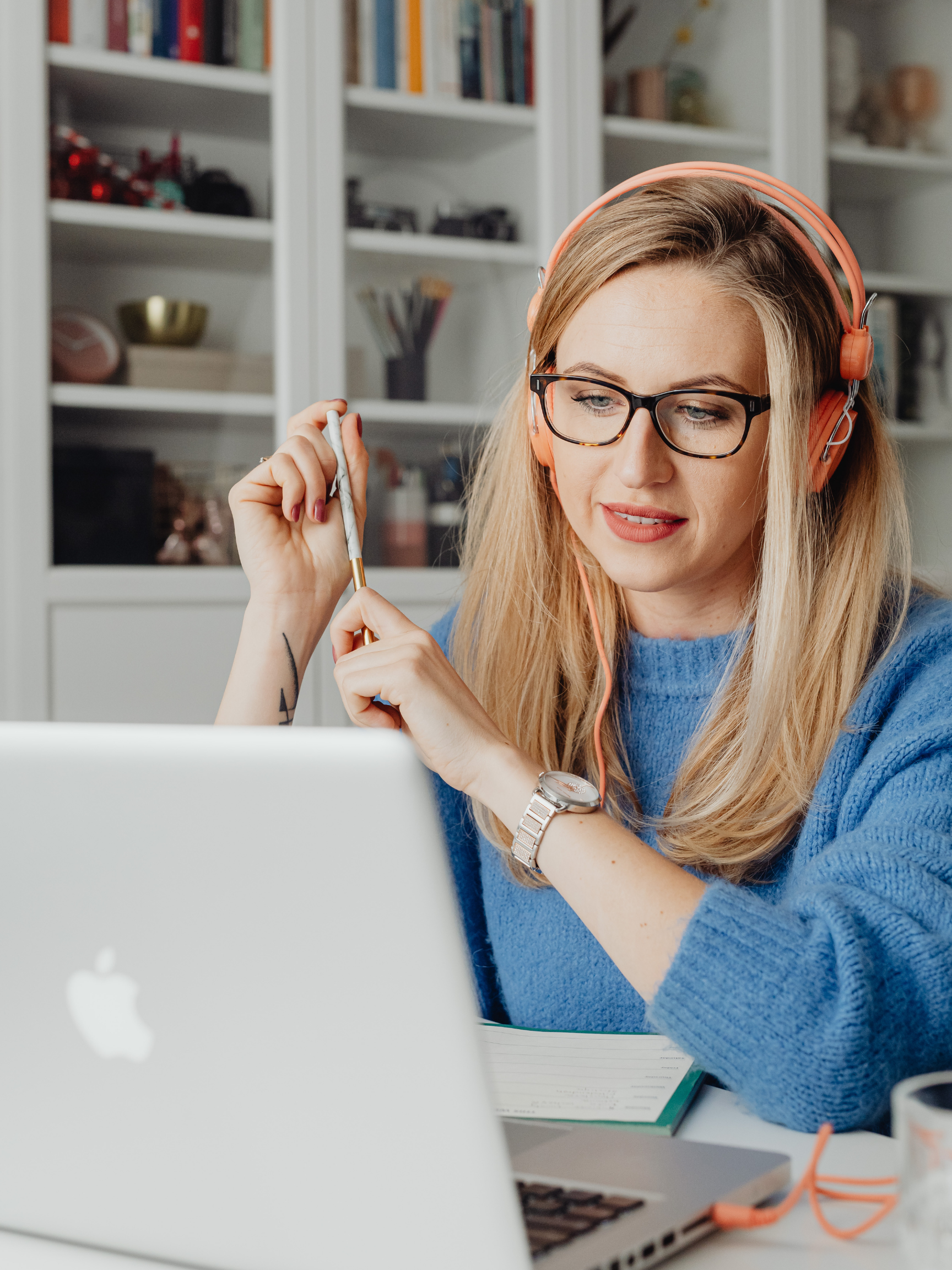 A woman with blonde hair, wearing glasses, a blue sweater, and pink headphones, is sitting at a desk with a laptop, holding a pen, and looking at her screen.