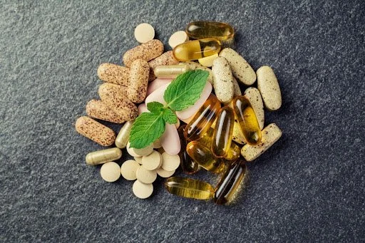 A collection of various pills and capsules, some with a green leaf placed among them, on a dark textured surface.