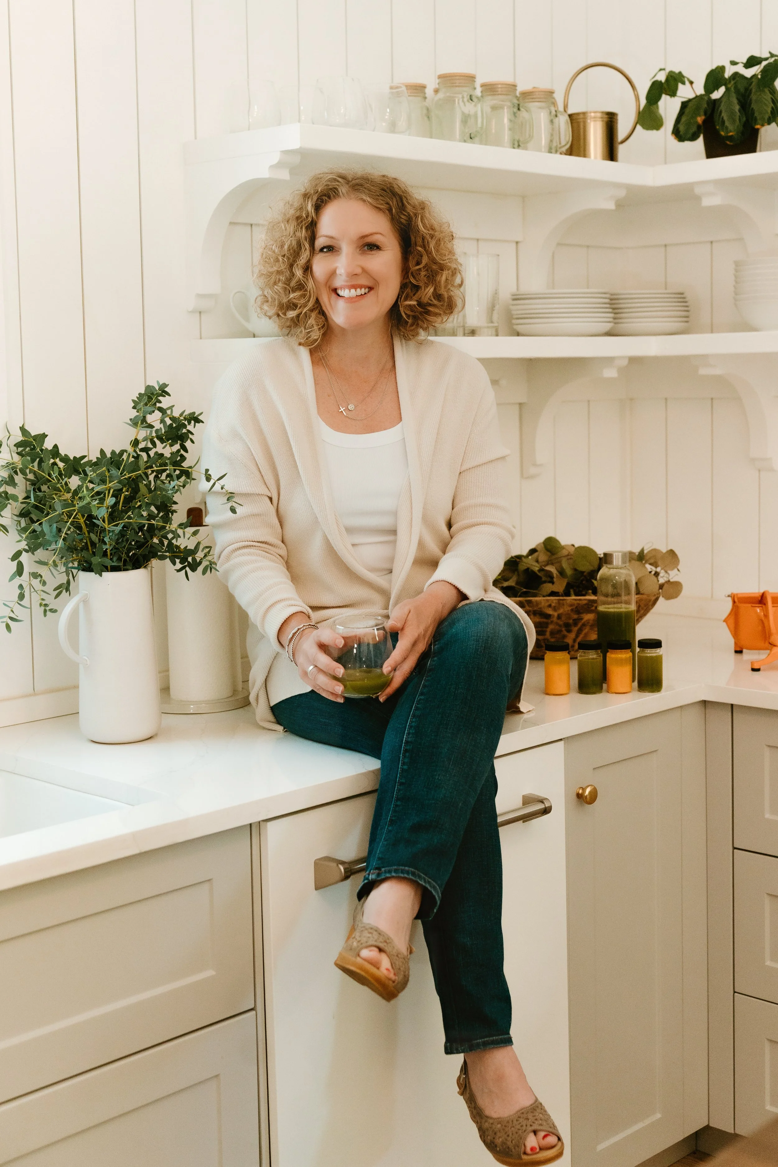 A woman with curly blonde hair sitting on a kitchen counter holding a glass of green smoothie, surrounded by plants, jars, and dishes in a bright, cozy kitchen.