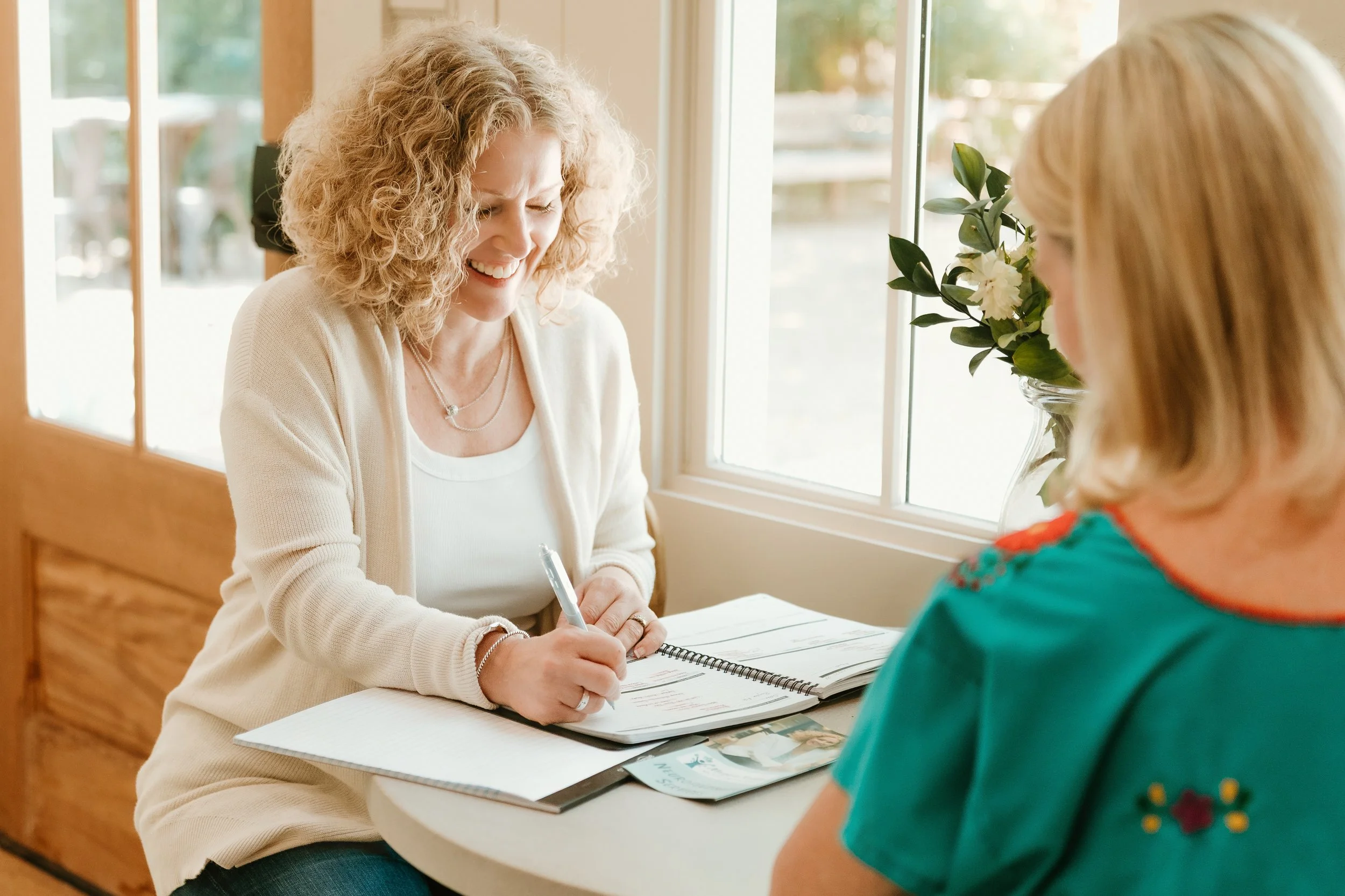 A woman with curly blonde hair smiling while sitting at a table, talking to another woman with blonde hair wearing a teal shirt, with a notebook, pen, and brochures in front of her, next to a window with a vase of white flowers.