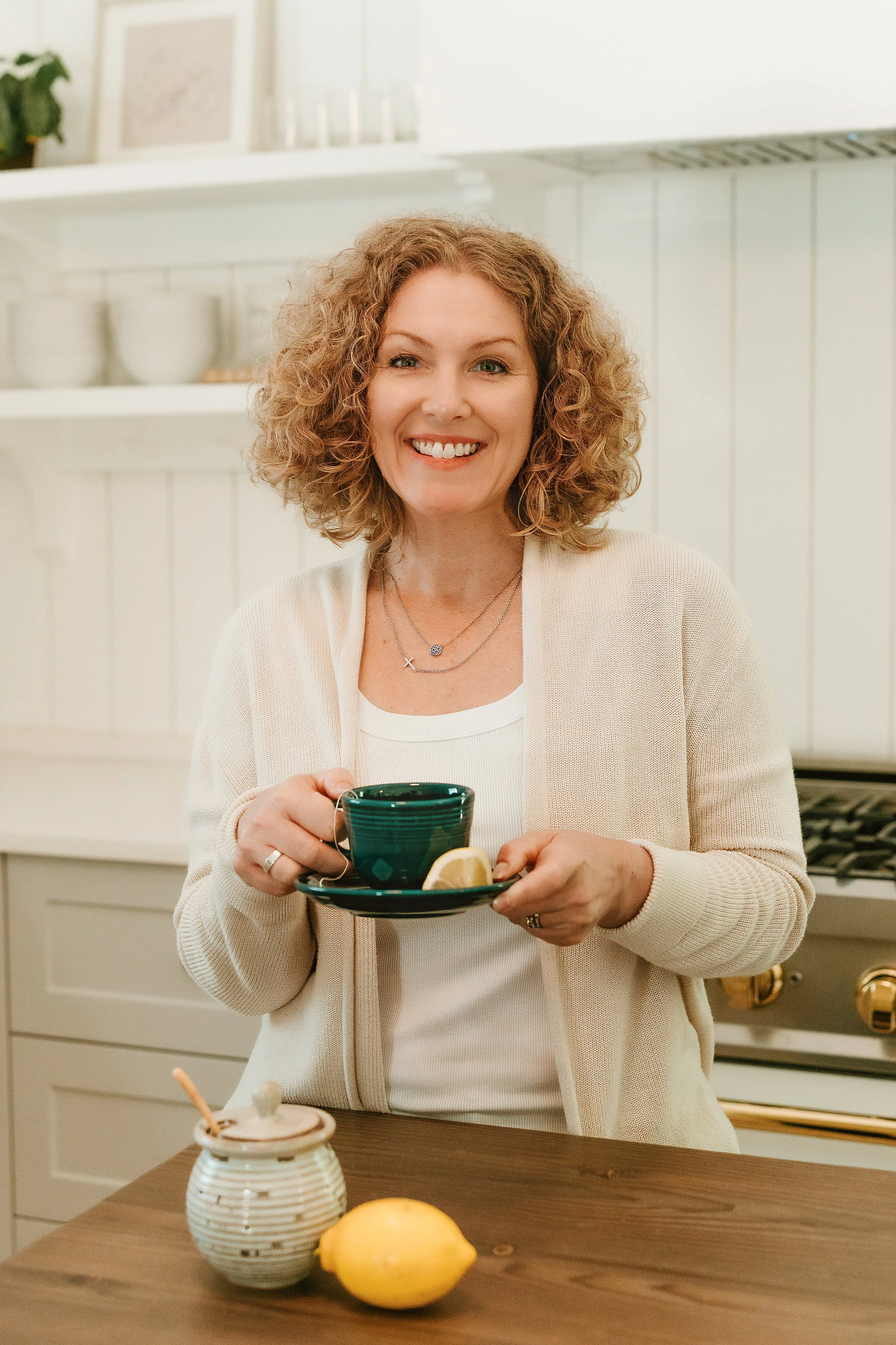 A smiling woman with curly hair holding a dark green cup and saucer with lemon, standing in a kitchen.