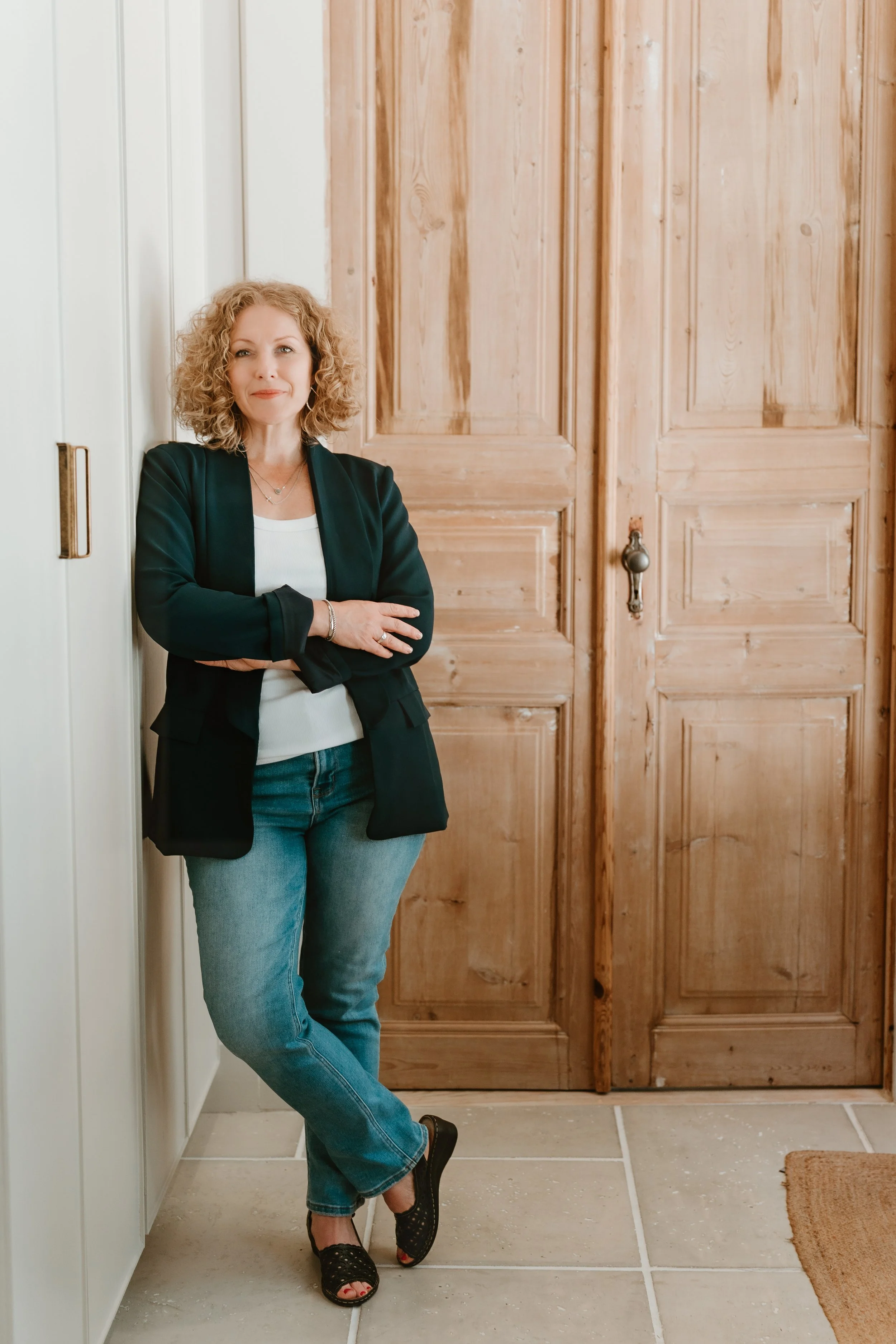 A woman with curly blonde hair standing in front of wooden doors in a home interior, wearing a black blazer, white top, blue jeans, and black open-toe shoes.