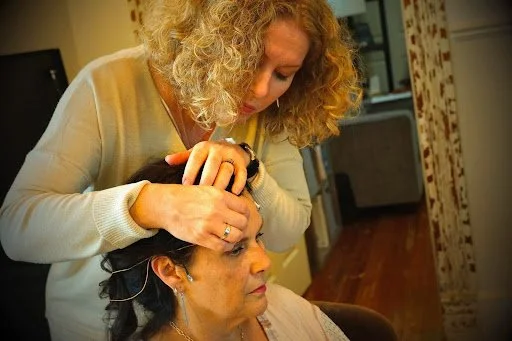 A woman with curly blonde hair styles the hair of another woman seated in a chair.