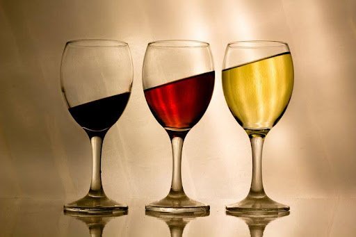 Three wine glasses, each filled with red, white, and rosé wine, placed on a reflective surface against a neutral background.