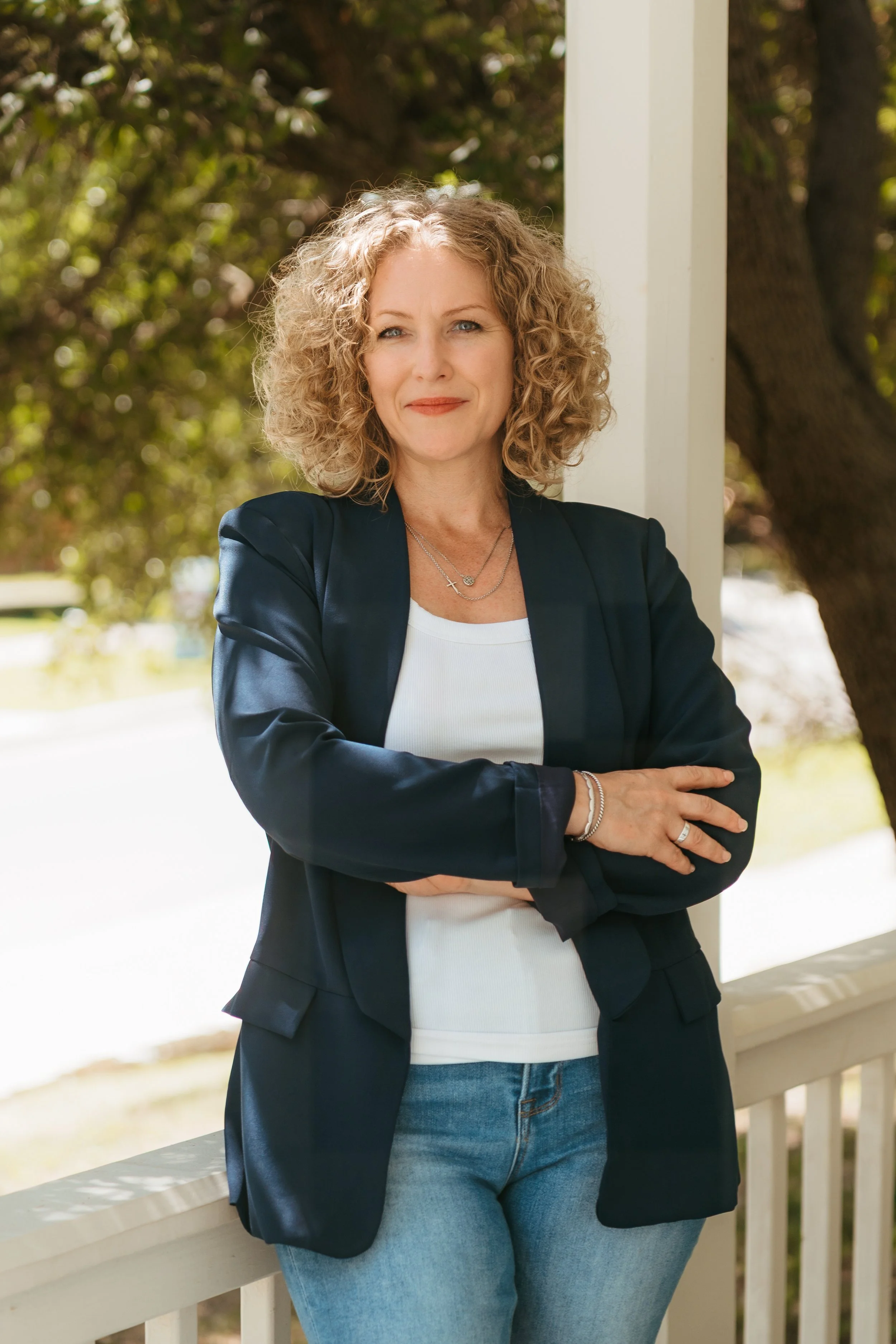 A woman with curly blonde hair standing outside on a porch with her arms crossed, wearing a dark blazer, white top, and jeans.