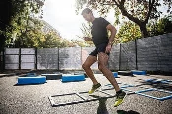 Man jumping over agility ladders on an outdoor fitness ground with trees and fencing in background.