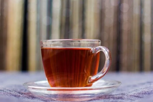 A clear glass coffee cup filled with dark hot tea on a matching glass saucer, placed on a purple cloth with a blurred background.