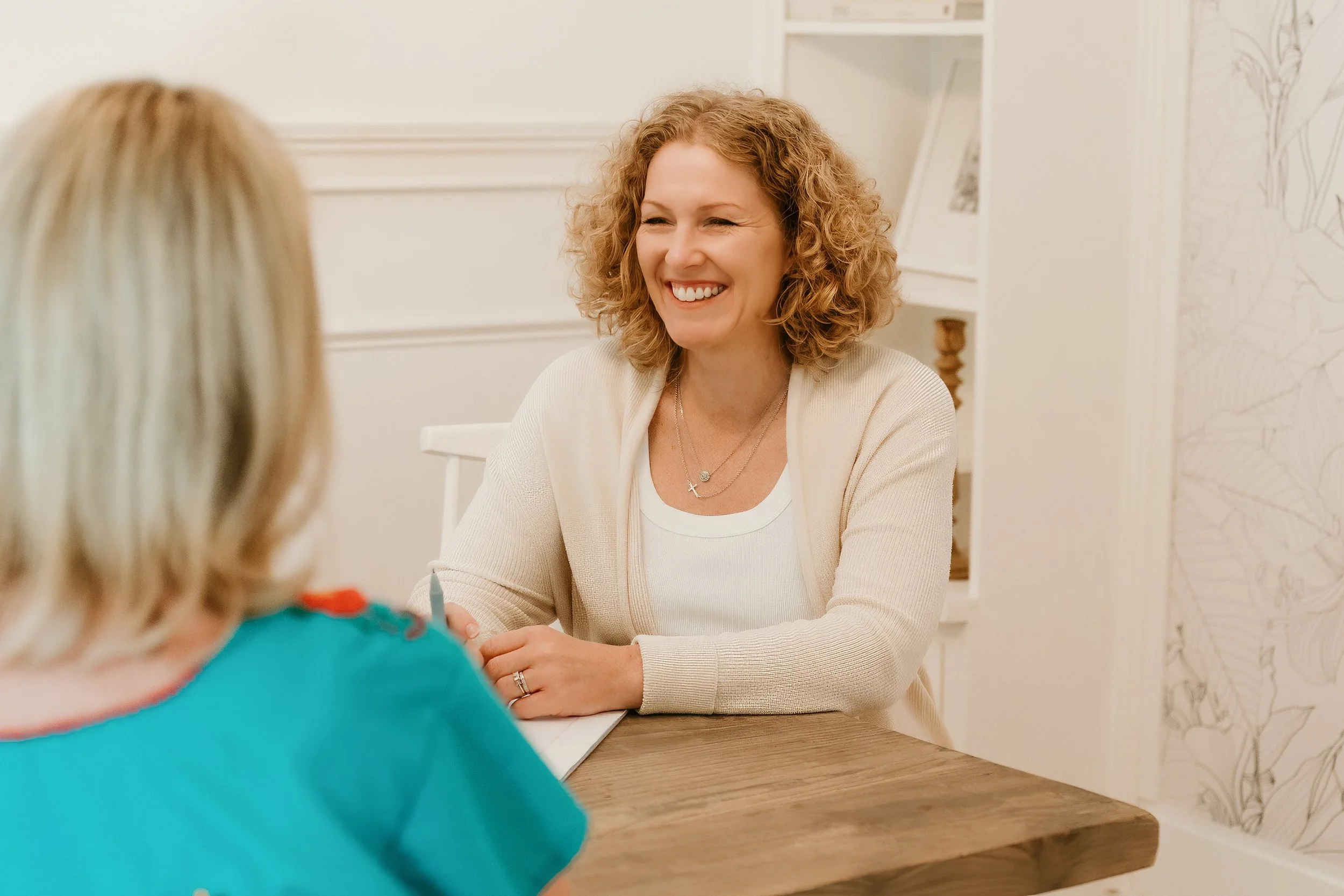 A woman with curly blonde hair smiling and talking to a person with blonde hair wearing a teal shirt, sitting at a wooden table in a bright room.