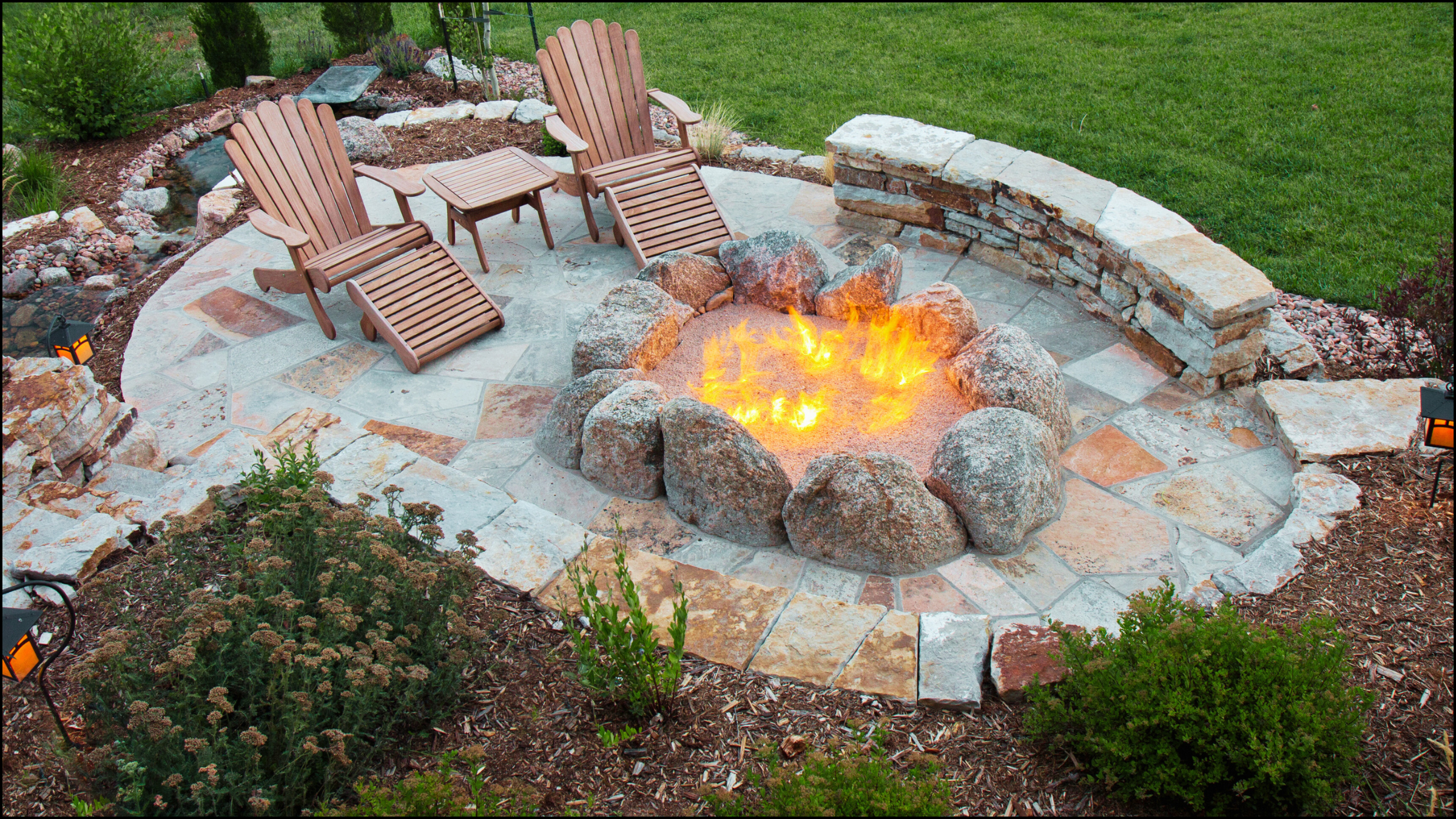 A circular outdoor seating area with two wooden Adirondack chairs, a side table, and a fire pit surrounded by large rocks. The area has stone pavers and is bordered by plants, shrubs, and a grassy lawn.