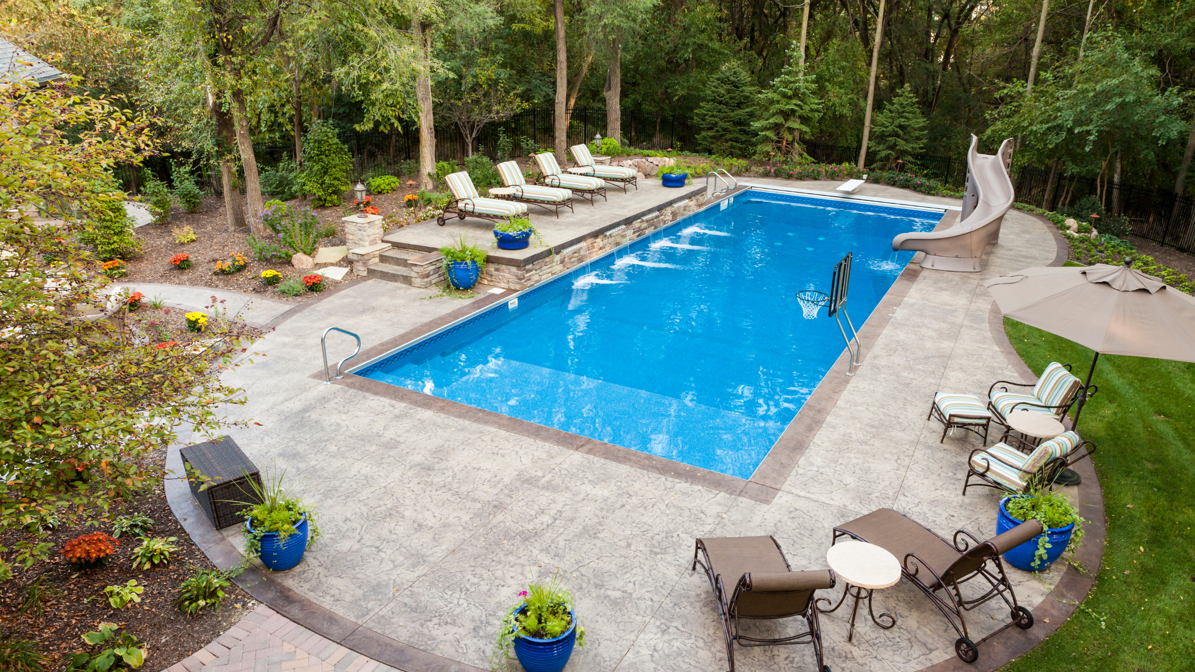 Backyard with a swimming pool surrounded by lounge chairs, potted plants, a large umbrella, and a slide, with trees and landscaping in the background.