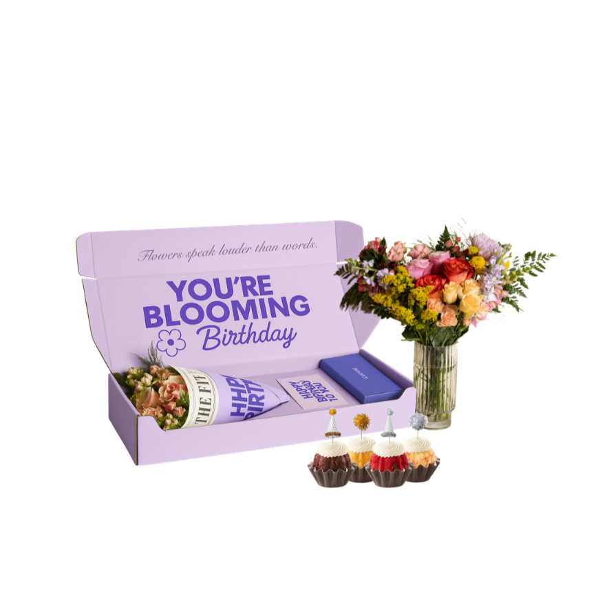 A birthday gift set with a purple box containing a bouquet of pink, peach, and yellow flowers along with a greeting card, a blue box, and a rolled-up paper reading 'The Fittest.' Beside the box is a vase of mixed flowers and three cupcakes with white, red, and chocolate frosting and decorative picks.