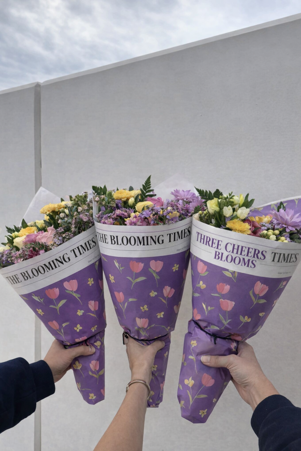 Three bouquets of colorful flowers wrapped in purple paper with floral patterns and newspaper-style labels, held by three different hands.
