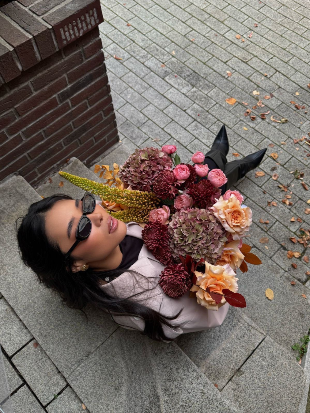 Woman with dark hair, wearing sunglasses and a light pink blazer, sitting on stone steps near a brick wall, holding a large bouquet of assorted pink, peach, and burgundy flowers.