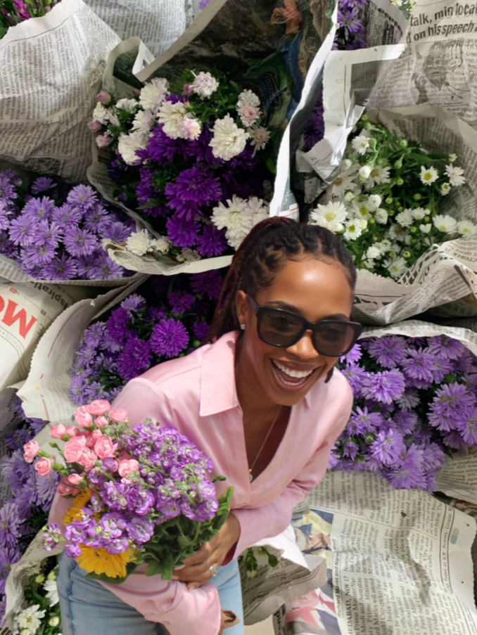 A woman with dreadlocks wearing sunglasses and a pink shirt, smiling and holding a colorful bouquet of pink, purple, and yellow flowers, standing among bouquets of purple and white flowers wrapped in newspaper.