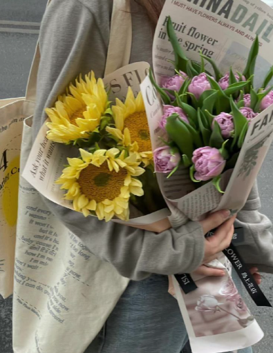 Person holding two bouquets of flowers, one with yellow sunflowers and the other with pink tulips, wrapped in newspaper.