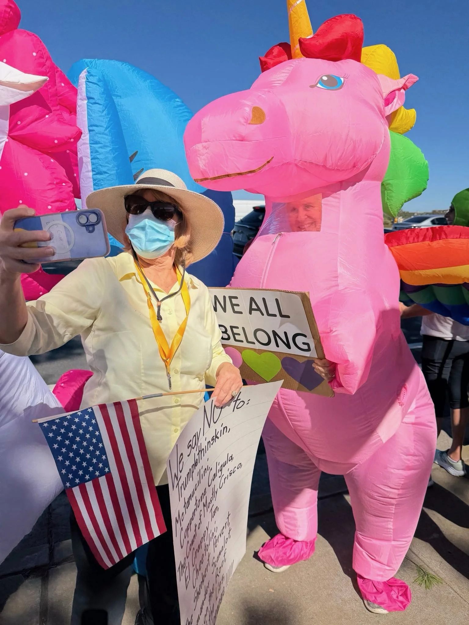 A woman wearing a straw hat, sunglasses, and a face mask takes a selfie with a person in a pink unicorn costume holding a sign that reads "WE ALL BELONG." The woman is holding an American flag and a poster with handwritten text. The scene takes place