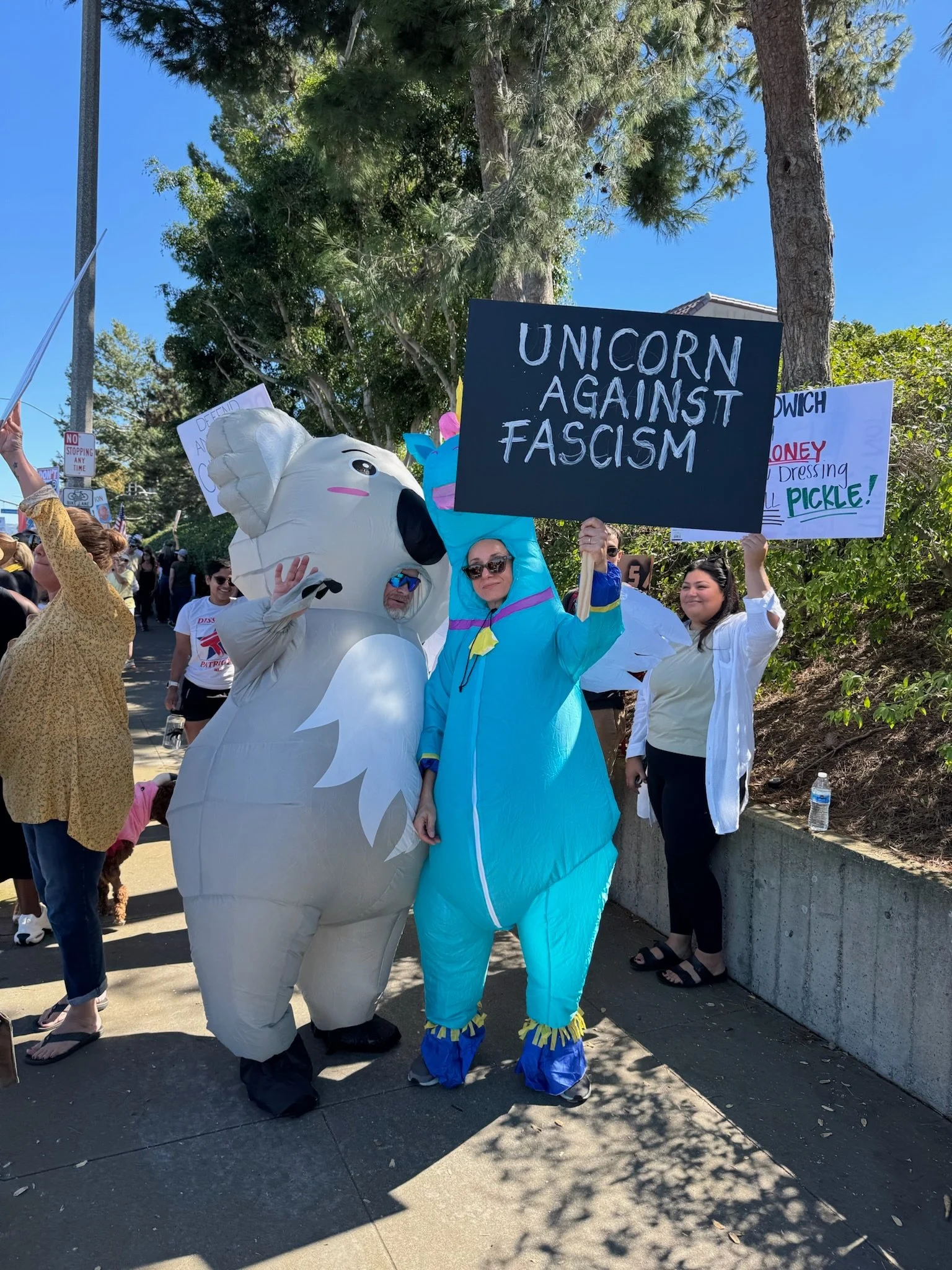 People at a protest or rally, one dressed in a koala costume and holding a sign that says 'Unicorn Against Fascism', others holding signs and standing among trees and sunlight.
