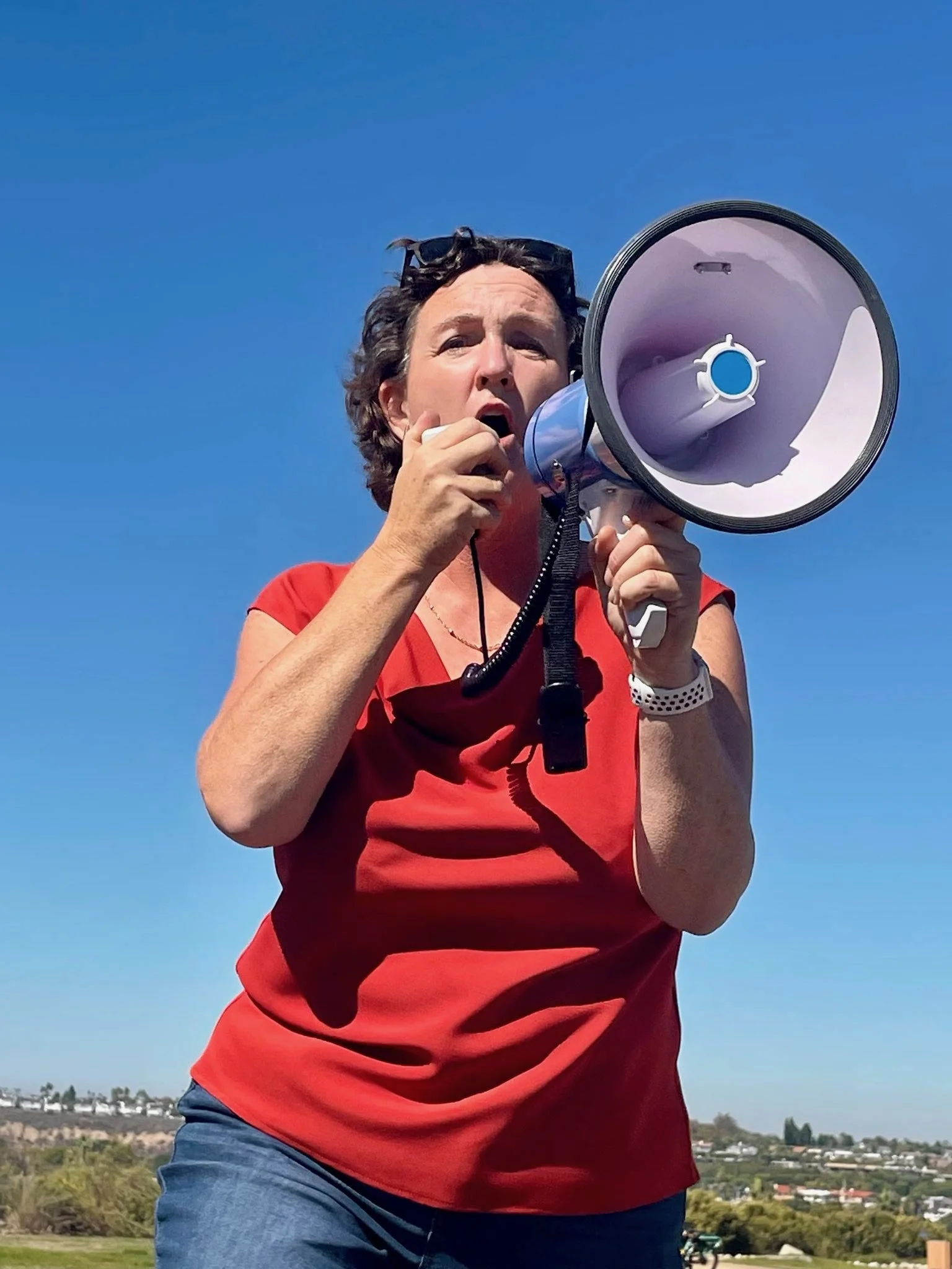 Woman in a red shirt speaking into a megaphone outdoors against a clear blue sky.
