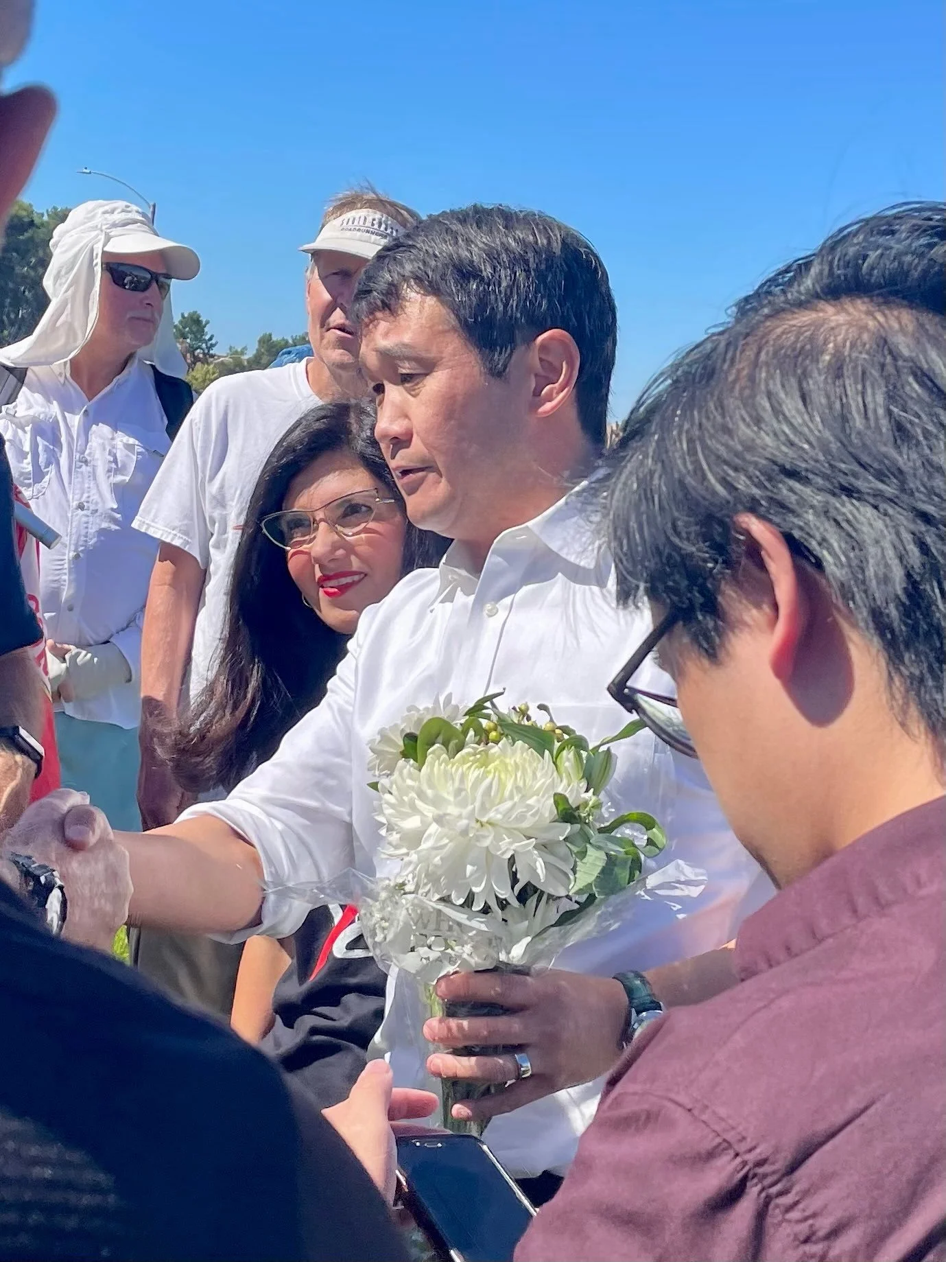 A man in a white shirt holding a bouquet of white flowers, surrounded by a group of people outdoors under a clear blue sky.