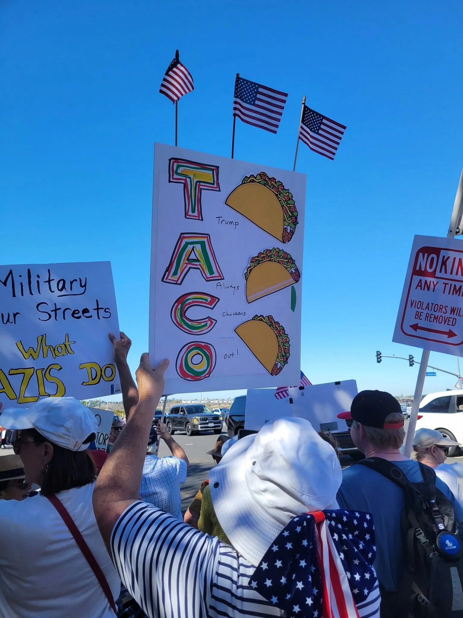 Protesters holding signs, one with a rainbow-colored 'TACO' and illustrations of tacos labeled with themes such as 'Trump,' 'Always chickens,' and 'out!'; others with messages against military presence and violations.