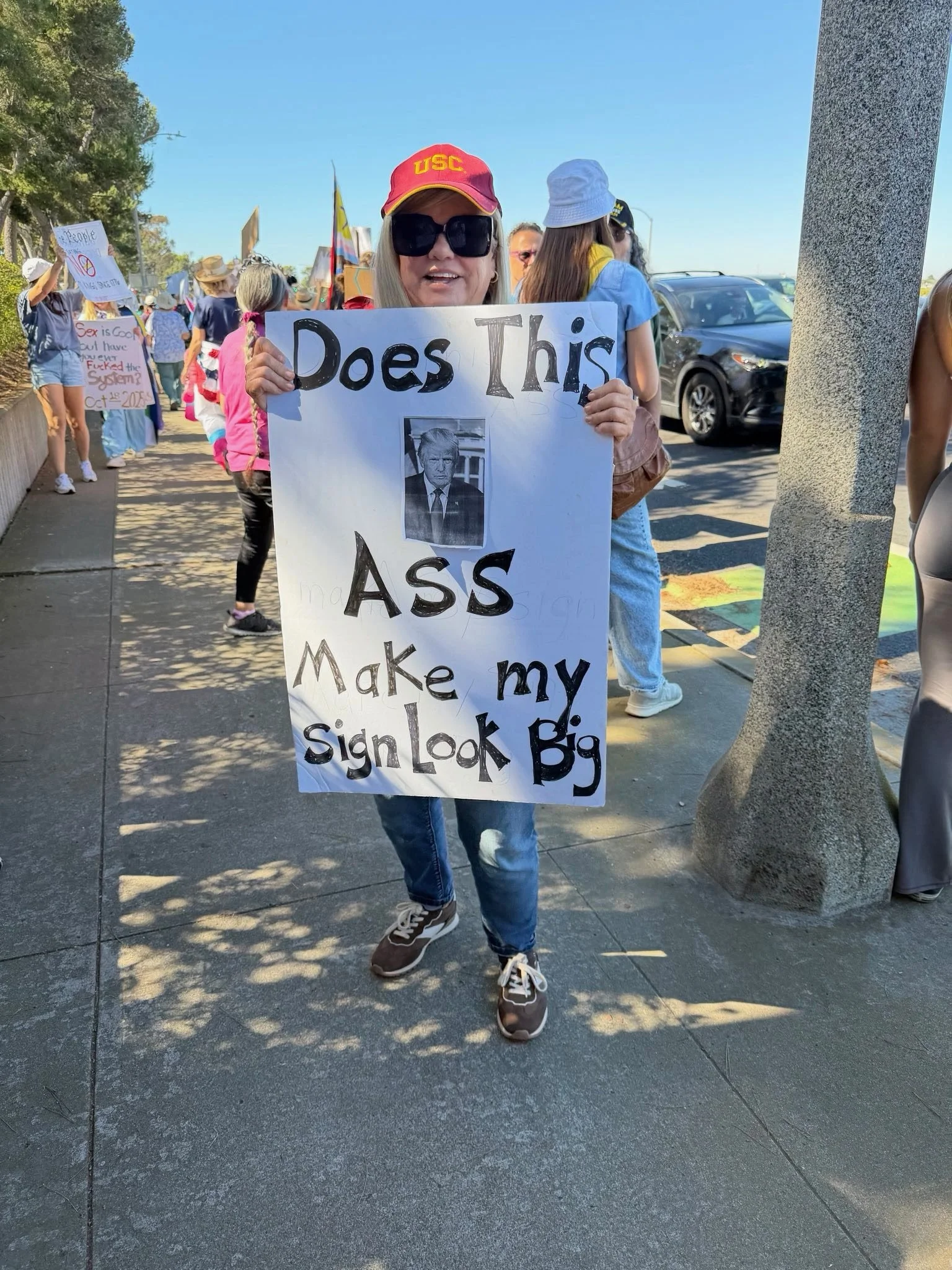 A protester holding a sign with a photo of Donald Trump and the words 'Does This Ass Make My Sign Look Big' at a rally, with other protesters in the background.