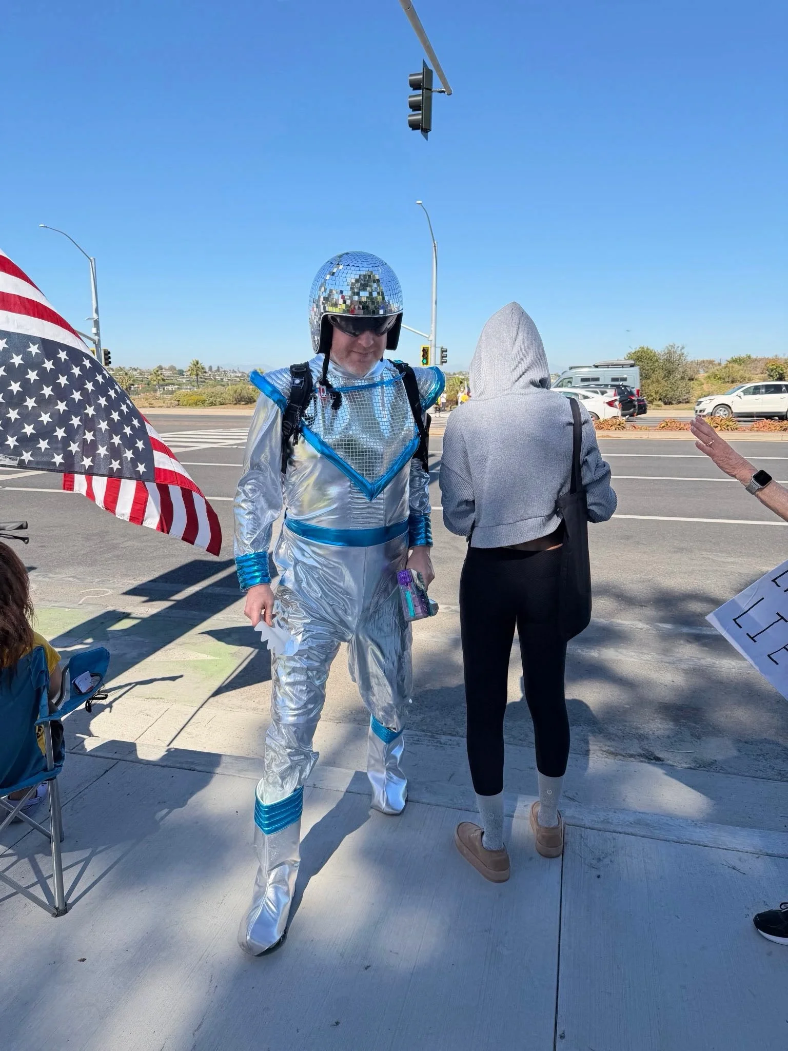 A man in a shiny, silver space-themed costume with a disco ball helmet standing on a sidewalk, talking to a woman with a gray hoodie and black leggings, near a corner store with American flags, cars, and traffic lights in the background.