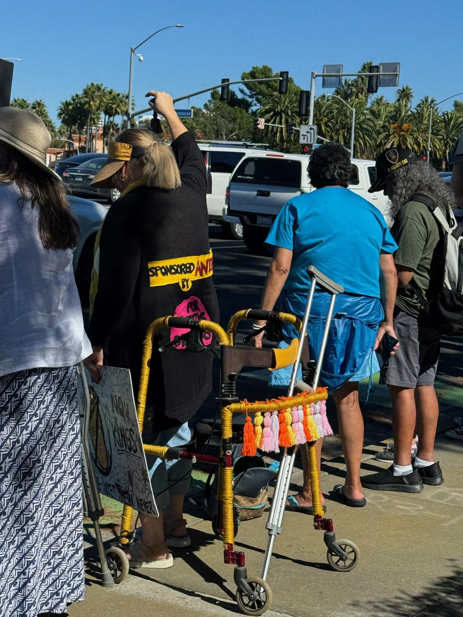 A group of people standing in line outdoors on a sidewalk, some using walkers, with palm trees, cars, and traffic lights in the background on a sunny day.