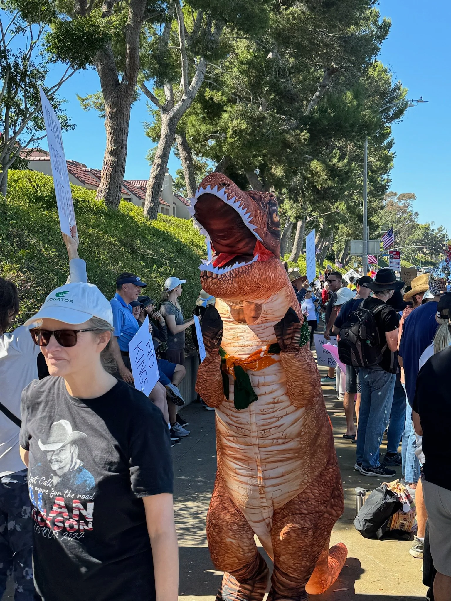 People participating in a march or protest, with some holding signs, and one person dressed in an inflatable T-Rex costume.