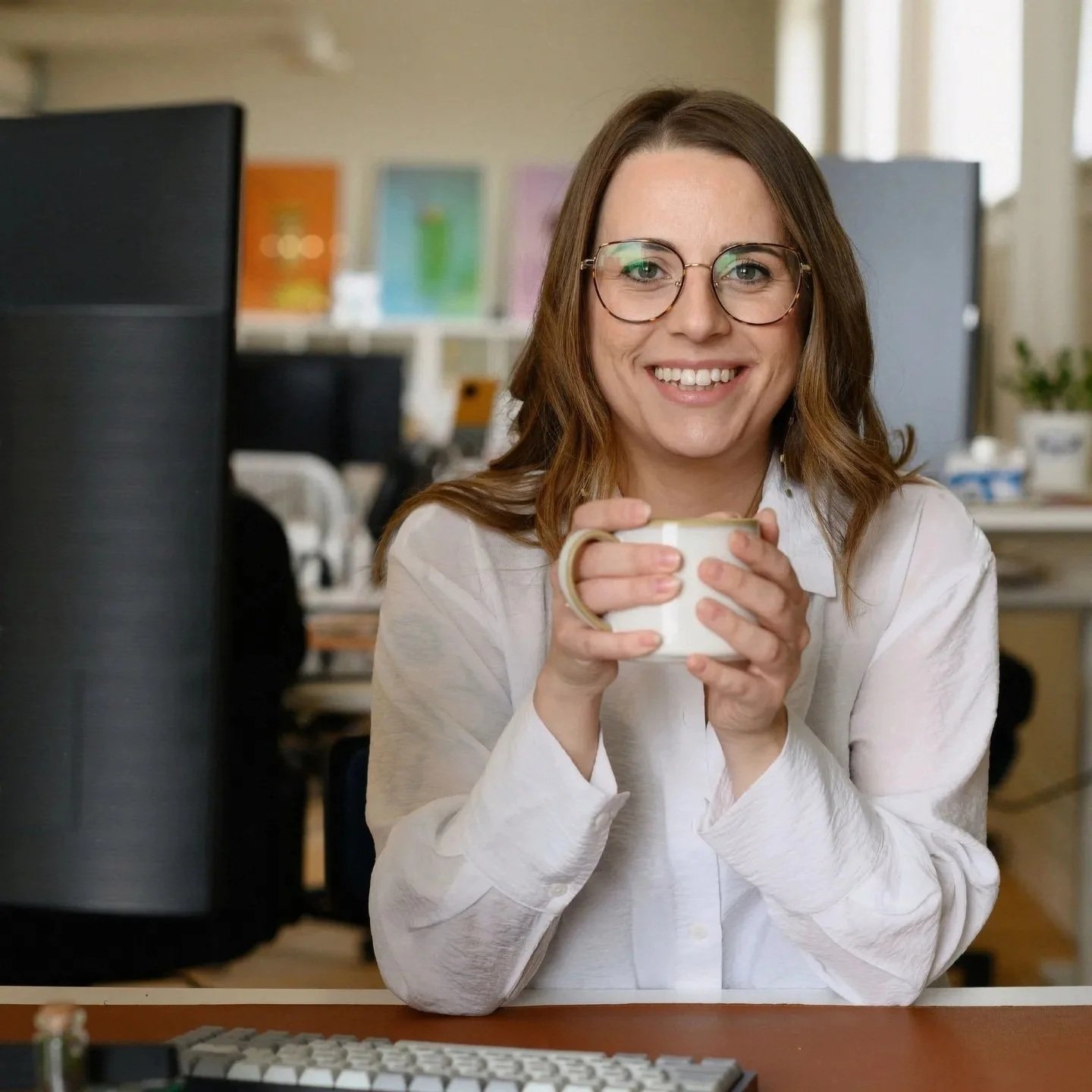 A woman with glasses smiling and holding a mug in an office setting.