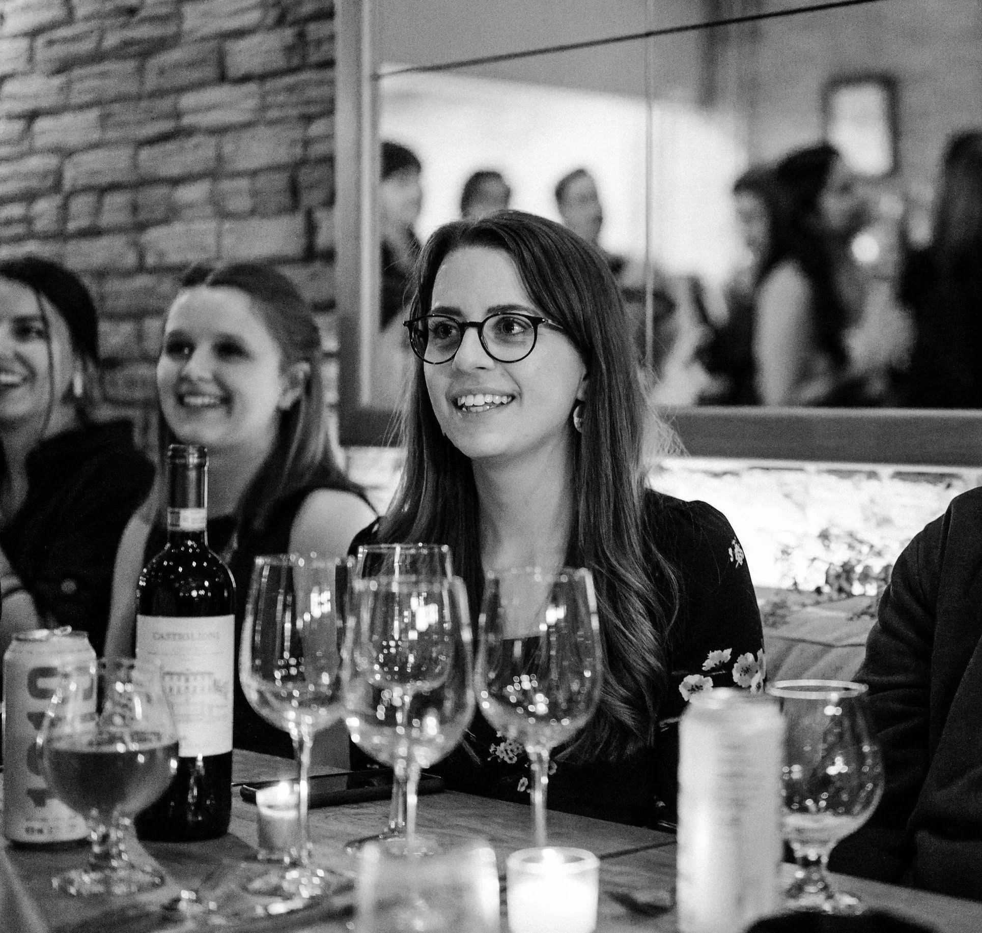 A woman with glasses and long hair smiling at a social gathering around a table with coworkers in the background.