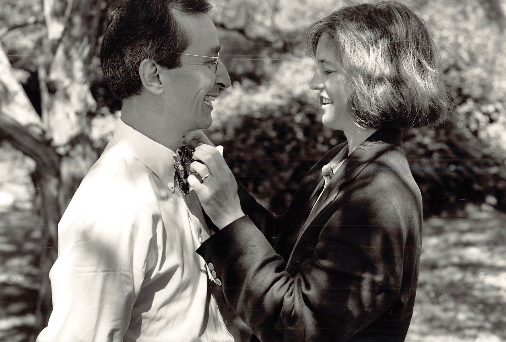 A black-and-white photo of a man wearing glasses and a woman with shoulder-length hair standing outdoors, smiling and gazing at each other, with the woman adjusting the man's tie.