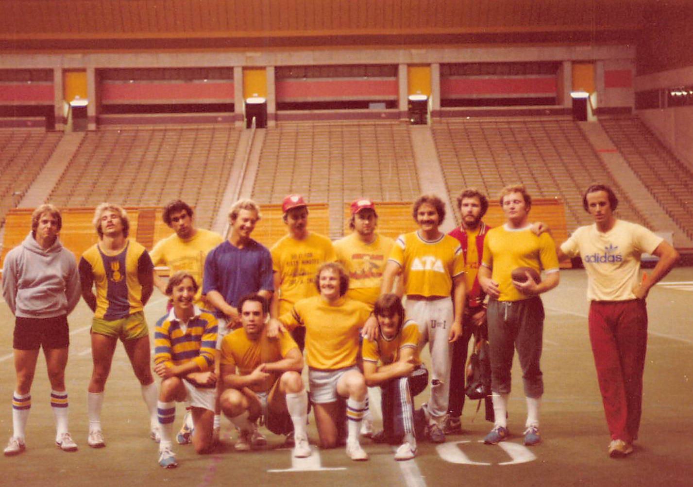 A group of fourteen young adults, mostly men, posing on an indoor football field. They are wearing athletic clothing in yellow and some are holding a football. The background shows a large, empty stadium with a high roof and tiered seating.