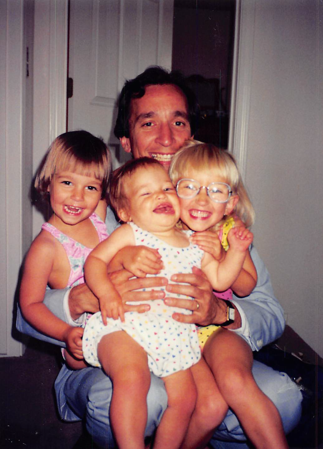Man sitting on a chair holding three young girls on his lap, all smiling and laughing indoors.