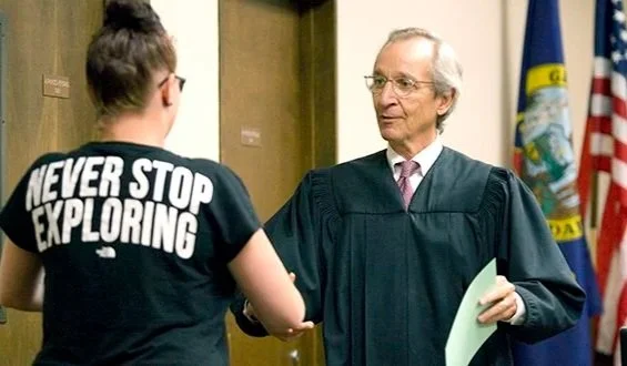 A judge shaking hands with a young woman in a courtroom, with American and state flags in the background.