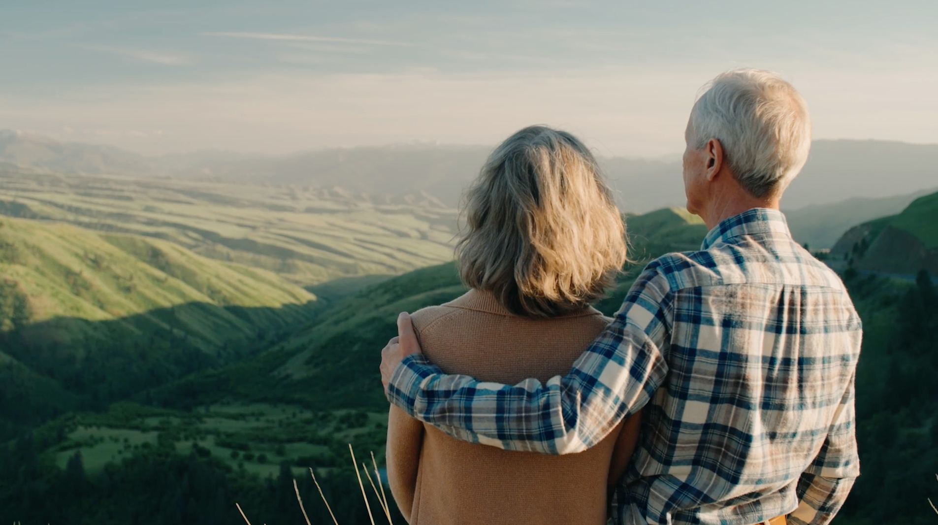 An elderly couple stands close together, facing away, overlooking lush green rolling hills under a partly cloudy sky.