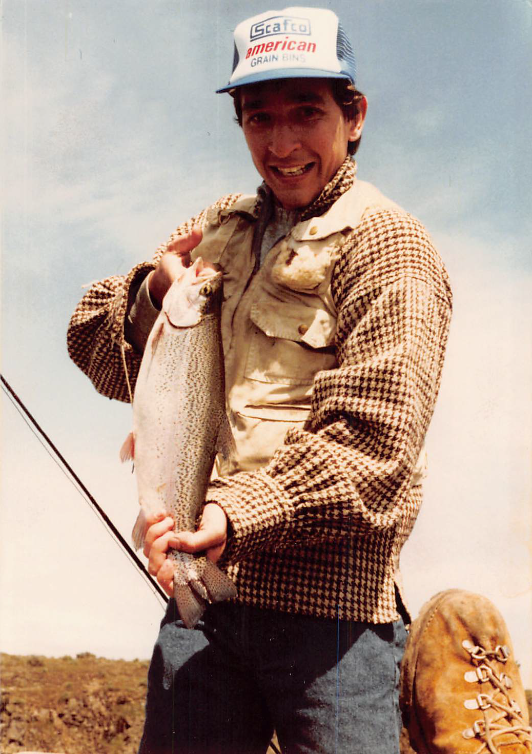 Man holding a large fish outdoors, wearing a plaid jacket, beige vest, and a baseball cap that reads 'Scafco American Grain Bins.'