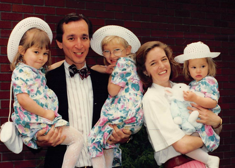 Family of five posing outdoors in front of a brick wall, with three young girls wearing matching floral dresses and sun hats, and two adults, a man in a tuxedo and a woman in a white blouse holding the youngest girl with a stuffed toy.