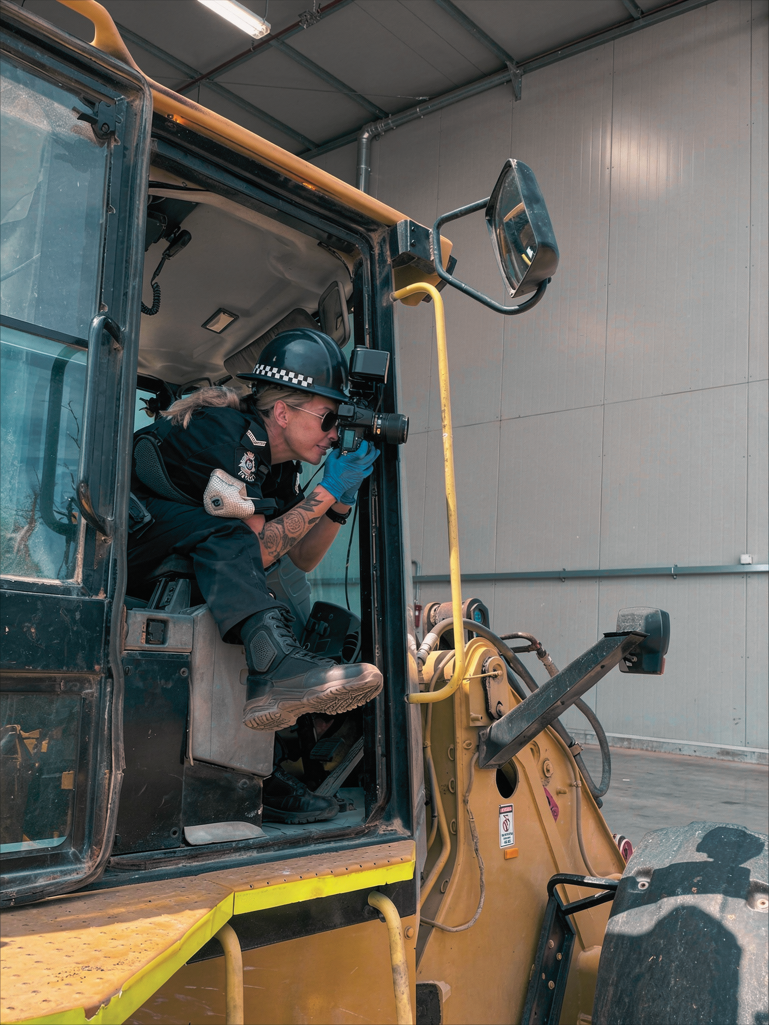 A police officer is sitting inside the cab of a construction vehicle, using a camera or binoculars to look through the window.