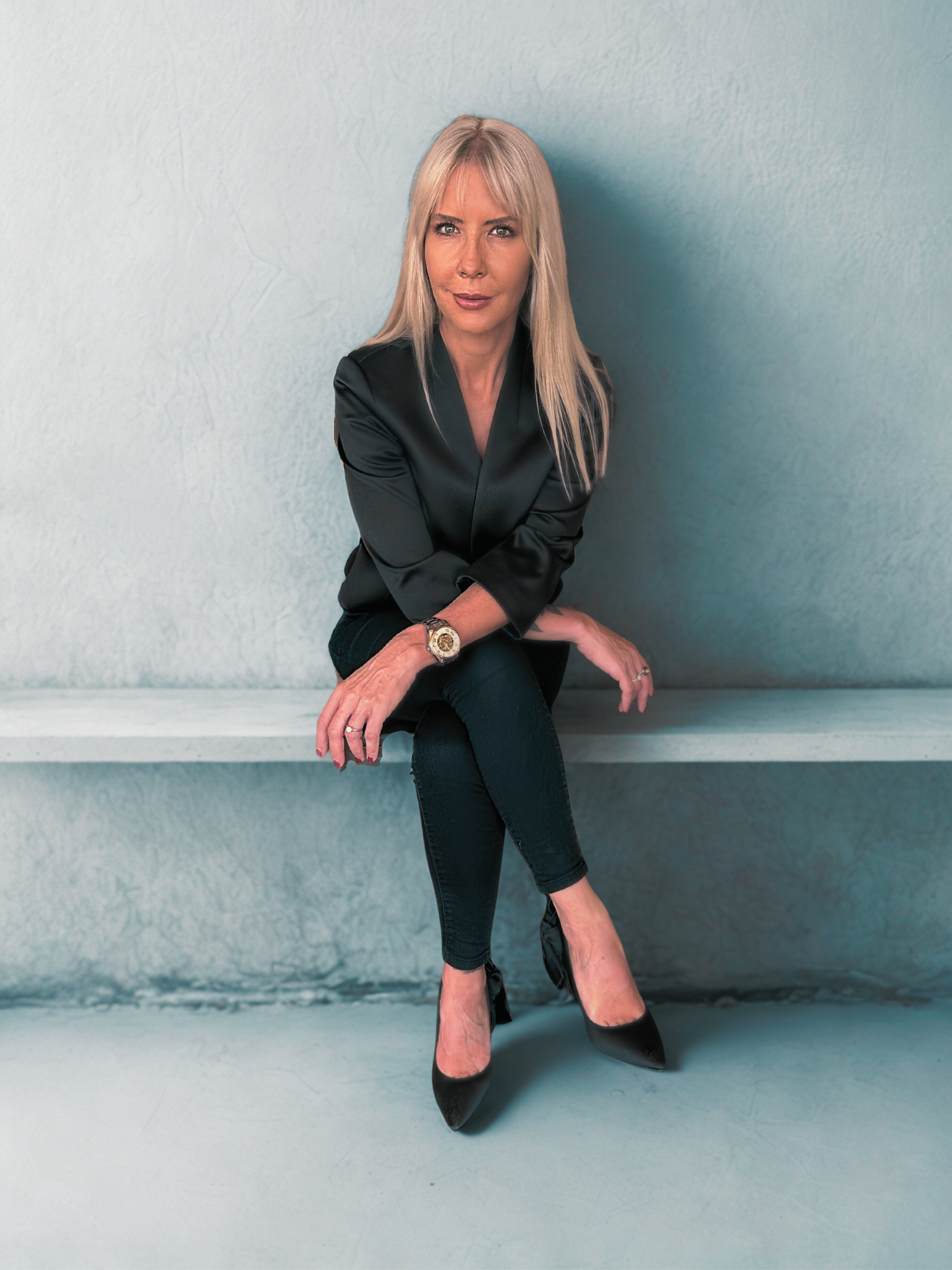 A woman with blonde hair wearing a black satin top and black pants sits on a white shelf against a plain light-colored wall.